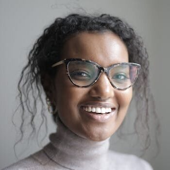 Young African American female in casual clothes and eyeglasses smiling and looking at camera while standing near blurred grey background